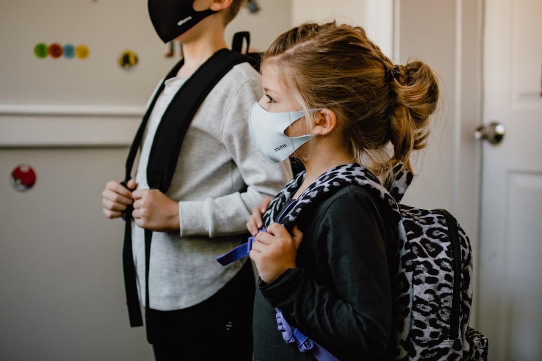 Två barn med ryggsäckar och ansiktsmasker i ett ljust rum med färgglada dekorationer.